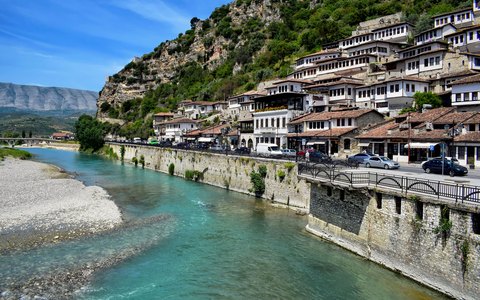 Reihen weißer osmanischer Häuser mit vielen Fenstern, die sich unter blauem Himmel einen grünen Hang über einem türkisfarbenen Fluss hinaufziehen