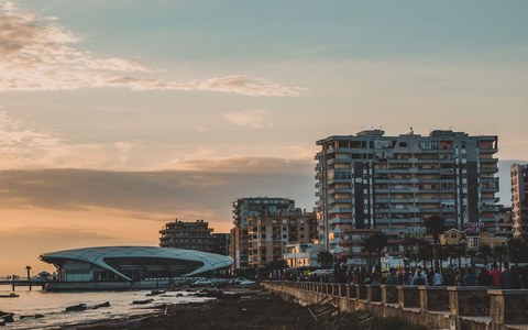 Die Uferpromenade von Durrës bei Sonnenuntergang, mit Wohnblöcken und Palmen sowie einem geschwungenen modernen Gebäude am Ufer