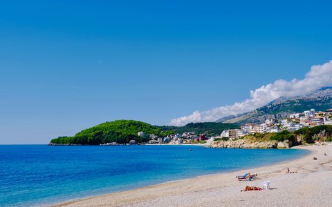 Ein Kiesstrand verläuft in einem Bogen entlang des klaren blauen Wassers zu den weißen Häusern von Himara und einer grünen Landzunge, dahinter Berge und ein tiefblauer Himmel