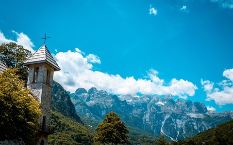 Ein kleiner steinerner Kirchturm vor schroffen grauen Bergen mit großen weißen Wolken am tiefblauen Himmel
