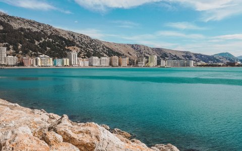 Klares türkisfarbenes Meer mit hellen Felsen im Vordergrund und einer Häuserzeile am Ufer unter kahlen Bergen, darüber blauer Himmel mit dünnen Wolken