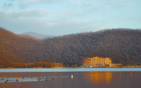 Nohur-See mit Enten auf dem Wasser, Hotel am gegenüberliegenden Ufer und bewaldeten Hügeln in Herbstfarben