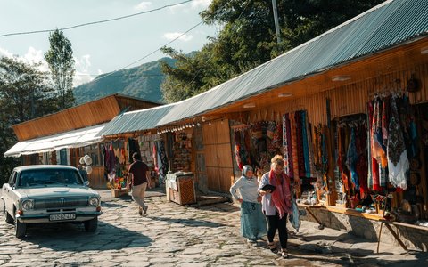 Kunsthandwerksstände mit Teppichen und Textilien an einer Kopfsteinpflasterstraße in Lahidsch, mit einem geparkten alten weißen Auto und vorbeigehenden Besuchern