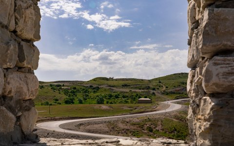 Blick zwischen alten Steinmauern hindurch auf eine gewundene Straße durch grüne Hügel bei Şamaxı unter blauem Himmel mit weißen Wolken