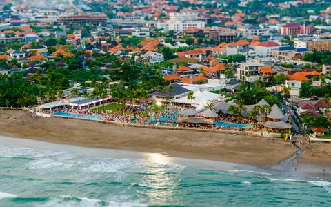 Luftaufnahme der Küste von Canggu mit Wellen, die auf den Sand rollen, einem Beachclub und der Stadt, die sich landeinwärts erstreckt