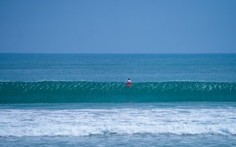 Ein einzelner Surfer paddelt am Strand von Kuta unter klarem Himmel durch türkisfarbene Wellen aufs Meer hinaus