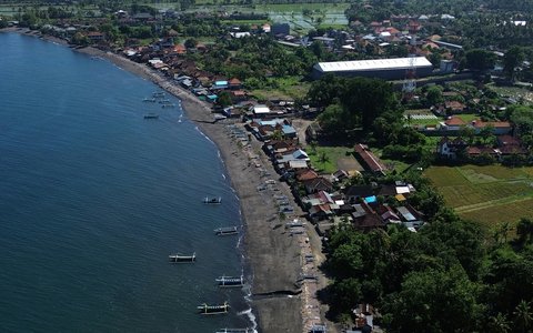 Luftaufnahme des dunklen Sandes am Lovina Beach mit Auslegerbooten im flachen Wasser und Dächern und Feldern des Dorfes dahinter