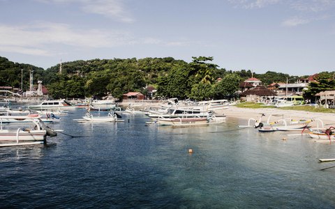 Auslegerboote liegen im Hafen von Padangbai unterhalb grüner Hügel vor Anker, darüber ein blauer Himmel mit Wolken
