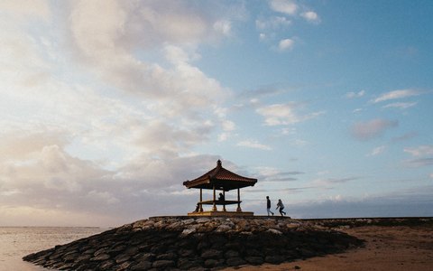 Ein traditioneller balinesischer Pavillon auf einer Steinzunge am ruhigen Meer bei Sanur unter zartblauem Himmel
