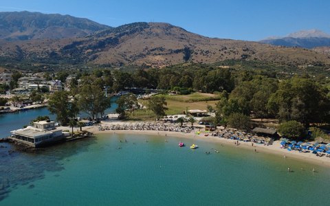 Luftaufnahme von Sandstrand und türkisfarbenem Wasser bei Georgioupoli, dahinter grüne Bäume und Berge