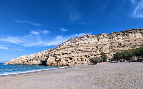 Kiesstrand unter hellen, von Höhlen durchzogenen Klippen bei Matala unter blauem Himmel mit weißen Wolken