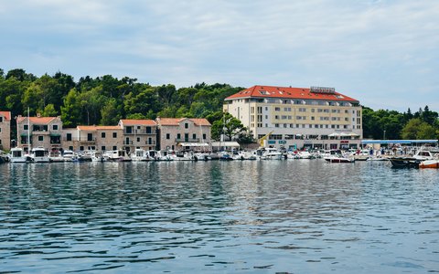 Boote in einem ruhigen Hafen vor Steingebäuden am Wasser und einem großen Hotel, mit grünen Bäumen und blauem Himmel