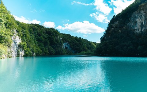 Blick auf einen türkisfarbenen See zwischen steilen bewaldeten Felswänden, mit weißen Wolken am blauen Himmel