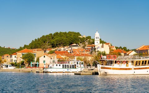 Boote liegen an der Uferpromenade von Skradin unterhalb eines Hügels mit Kirchturm und rot gedeckten Häusern unter klarem blauem Himmel