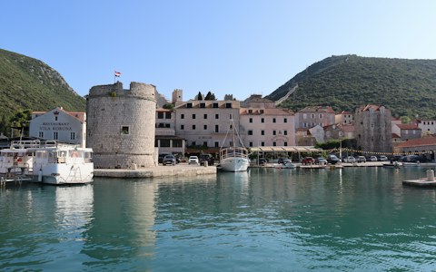 Runder Steinturm, Boote und pastellfarbene Häuser am Hafen von Mali Ston, dahinter grüne Hügel und alte Stadtmauern