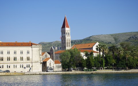 Unter blauem Himmel stehen Steingebäude und der Glockenturm der Kathedrale an der Uferpromenade von Trogir, dahinter grüne Hügel