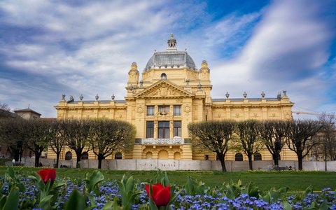Blick auf einen gelben Jugendstil-Pavillon mit Glaskuppel hinter Frühlingsblumenbeeten und Bäumen