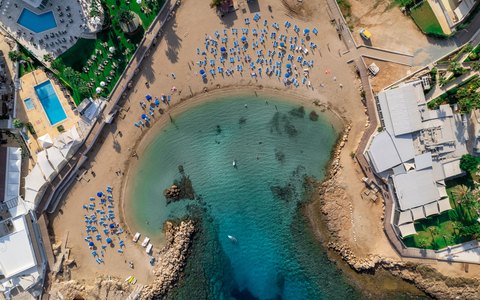 Blick aus der Vogelperspektive auf eine kleine Sandbucht mit klarem türkisfarbenem Wasser und Sonnenliegen entlang des Ufers