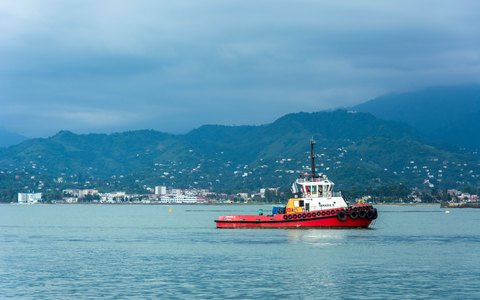 Blick auf einen roten Schlepper, der den Hafen von Batumi durchquert, mit der Stadt am Ufer und grünen Bergen im Hintergrund