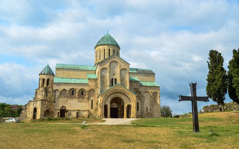 Steinerne Bagrati-Kathedrale auf einem grünen Hügel mit hoher Zypresse unter blauem Himmel und weißen Wolken
