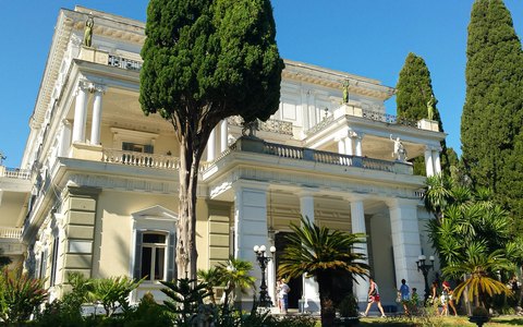 Der weiße neoklassizistische Achilleion-Palast hinter hohen Zypressen und Palmen unter blauem Himmel