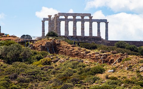 Marmorsäulen des Poseidontempels auf einer grünen felsigen Anhöhe unter blauem Himmel mit weißen Wolken