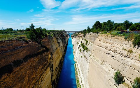 Der schmale Kanal von Korinth tief zwischen steilen Felswänden, darüber eine Brücke unter blauem Himmel mit weißen Wolken