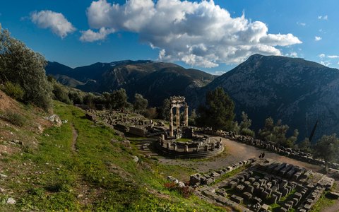 Die antiken Ruinen von Delphi an einem grünen Hang unterhalb der Berge, unter blauem Himmel mit weißen Wolken