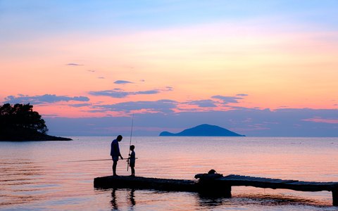 Vater und Sohn beim Angeln auf einem kleinen Steg in Neos Marmaras, als Silhouetten vor einem rosa Sonnenuntergang über Meer und Inseln
