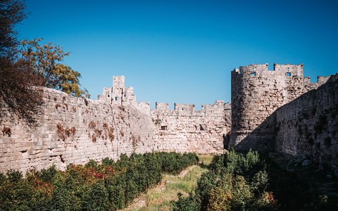 Blick auf die massiven Steinmauern und einen runden Turm der mittelalterlichen Stadt Rhodos, davor Pflanzen unter blauem Himmel