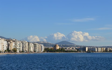 Blick über blaues Wasser auf Thessalonikis Ufer mit dem Weißen Turm und Wohnblocks, dahinter Berge und weiße Wolken