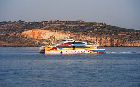 Eine bunt bemalte Gozo-Fähre überquert die blaue Meerenge und fährt dabei an hellen Kalksteinklippen vorbei