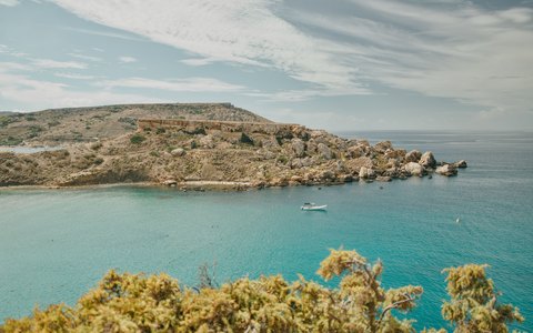 Blick auf das türkisfarbene Wasser unterhalb heller Kalksteinklippen an der Golden Bay, mit grünem Gestrüpp im Vordergrund und bewölktem Himmel