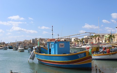 Blick auf die bemalten hölzernen Fischerboote, die im Hafen von Marsaxlokk vertäut liegen, dahinter die Uferpromenade des Dorfes unter blauem Himmel mit weißen Wolken