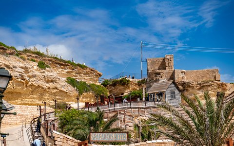 Blick auf die Holzhäuser von Popeye Village an einer felsigen Küste über dem Meer, mit Palmen und blauem Himmel mit weißen Wolken