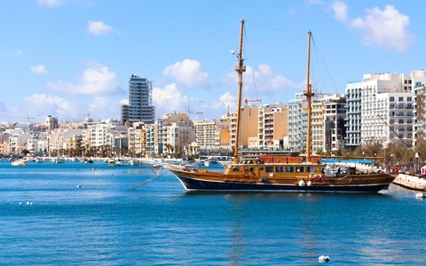 Hölzernes Segelschiff, das in ruhigem blauem Wasser vor der Uferpromenade von Sliema unter blauem Himmel vertäut liegt