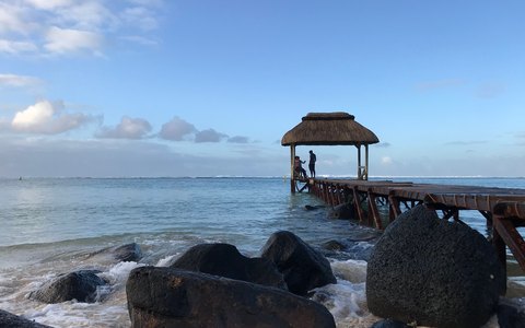 Ein strohgedeckter Pavillon am Ende eines Holzstegs über dem Meer in Bel Ombre, davor dunkle Felsen und weiße Wolken am blauen Himmel