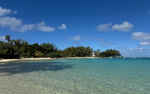 Blick auf das klare türkisfarbene Flachwasser und einen von Bäumen gesäumten Sandstrand in Blue Bay unter blauem Himmel mit weißen Wolken