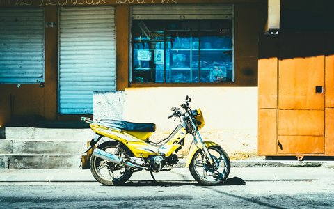 Gelbes Motorrad, das auf einer sonnigen Straße vor einem Geschäft mit grauen Metallrollläden und orangefarbenen Tafeln in Mahébourg geparkt ist