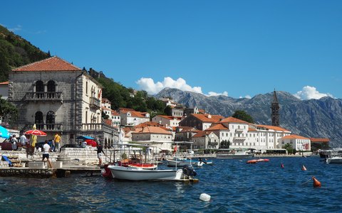 Blick auf die Steinhäuser und Boote an der Uferpromenade von Perast, mit Bergen auf der anderen Seite der Bucht und weißen Wolken am blauen Himmel