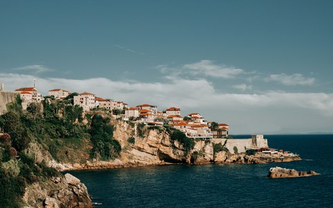 Unter blassblauem Himmel stehen alte Steingebäude auf einer grünen Klippe über der blauen Adria