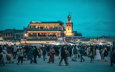 Menschenmenge auf dem Platz Djemaa el-Fna in der Dämmerung vor beleuchteten Caféterrassen unter einem tiefblauen Abendhimmel