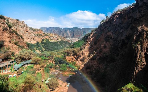 Blick auf eine grüne Schlucht mit roten Felswänden und einem Regenbogen in der Gischt der Ouzoud-Wasserfälle unter klarem blauem Himmel