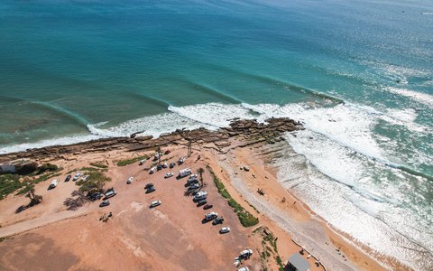Luftbild der Küste von Taghazout mit brechenden Wellen an einer sandigen Landzunge und Vans, die oben auf der Klippe geparkt sind