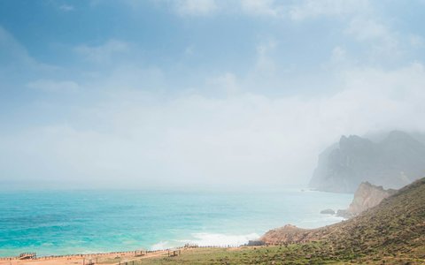 Türkisfarbenes Meer und ein geschwungener Strand bei Mughsail, mit Dunst über den Küstenklippen und grünen Hängen im Vordergrund