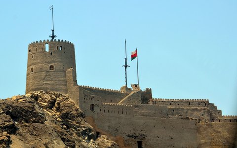 Unter klarem blauem Himmel das steinerne Fort Mutrah mit seinem runden Turm und einer Flagge auf einem felsigen Hügel