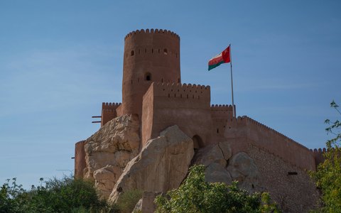 Der runde Steinturm des Nakhal-Forts mit der omanischen Flagge auf einer felsigen Hügelkuppe unter tiefblauem Himmel