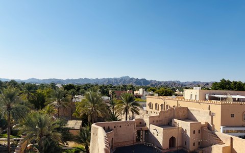 Blick auf das Fort Nizwa und die Palmenoase ringsum, dahinter das Hadschar-Gebirge, unter klarem blauem Himmel
