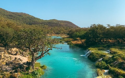 Ein türkisfarbener Bach mit kleinen Wasserfällen fließt zwischen grünen Bäumen und Felsen, unter bewaldeten Hügeln und blauem Himmel