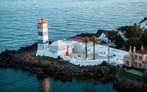 Luftaufnahme des Leuchtturms Santa Marta und weißer Gebäude am felsigen Ufer von Cascais, neben dem blauen Meer